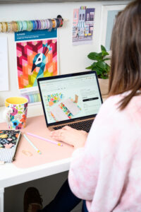 Woman with brown hair wearing pink sweater sitting at a colorful desk typing on a laptop