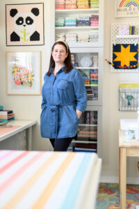 Woman wearing a blue shirt dress with brown hair and headband standing in a colorful sewing room
