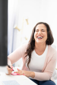 Woman with long dark brown hair is sitting at a white desk holding a pen and paper. She has her eyes closed and is laughing. She is wearing a white tank top with a light pink cardigan. The background is a mainly white offie and is out of focus.