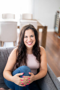 Woman with long dark brown hair is sitting in a living room on a light grey couch. She has her legs curled up and is smiling at the camera. There is a dining room table and white chairs out of focus in the background.