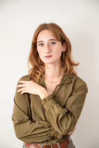 Woman with long red wavy hair standing against white wall. She's wearing a long sleeve olive colored silk top and gold paper clip chain jewellery.