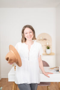 Woman is standing in front of a white desk. She is holding a light brown hat and wearing a white flowy short sleeved top with eyelet buttons on the front. She has shoulder length dark blonde har and is smiling at the camera. Behind her is a wooden shelf that is slightly out of focus and a white wall.