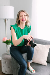 Woman sitting on grey couch laughing. She's wearing a green top, jeans and floral Keds. She's holding a camera.