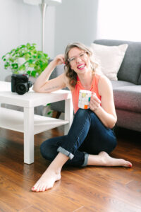 Woman sitting on the floor leaning on a white coffee table. She's holding a mug on her knee and she's laughing