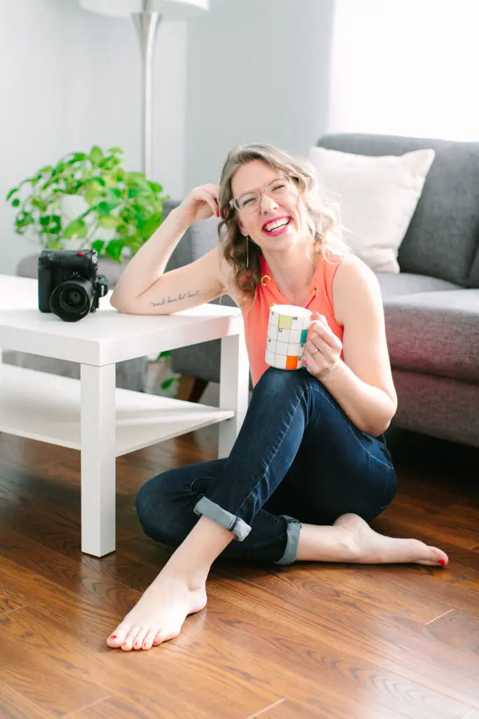 Amy sitting on the floor leaning on a white coffee table. She's holding a mug on her knee and she's laughing.