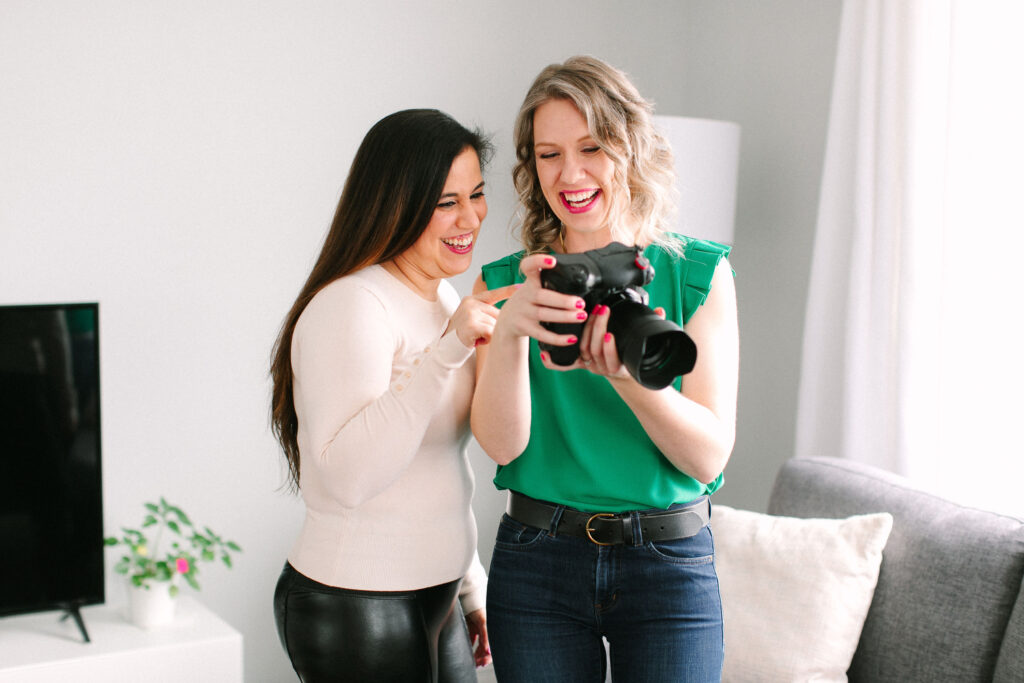 Amy is standing beside a client wearing a green shirt. She is smiling and appears to be showing her client the images on the back of her camera screen.